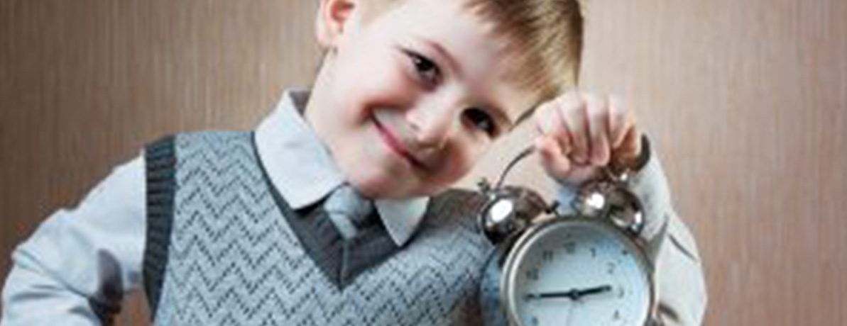 A young boy is smiling, wearing a sweater vest with a tie while holding up a clock