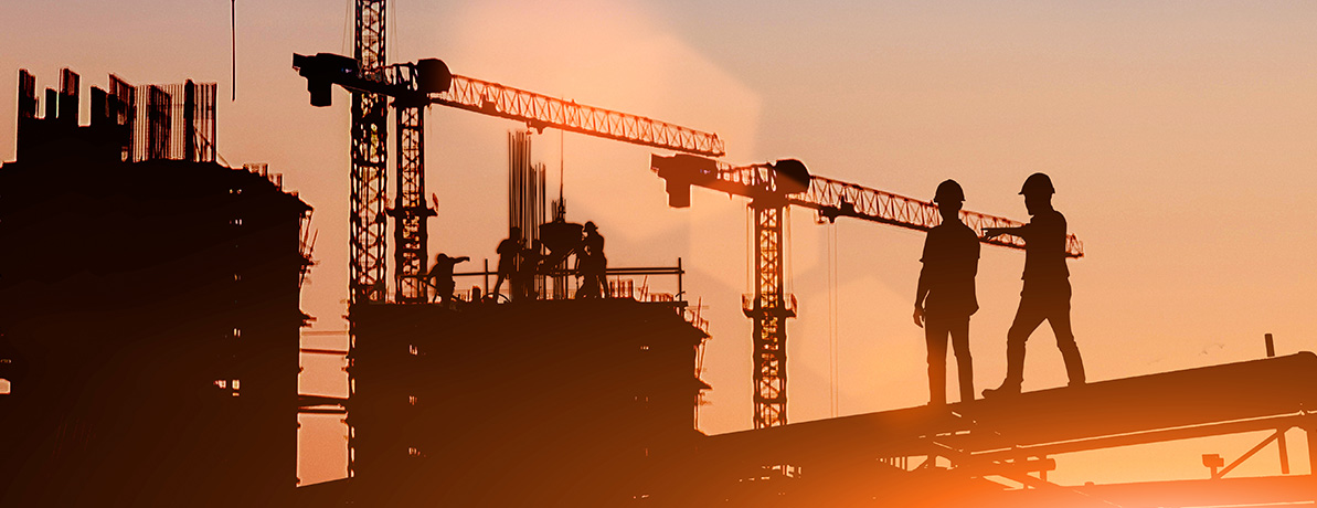 Silhouette de 2 constructeurs signalant du doigt d’autres ouvriers travaillant sur un chantier, avec des grues en arrière-plan. Soleil en train de se coucher dans le coin.